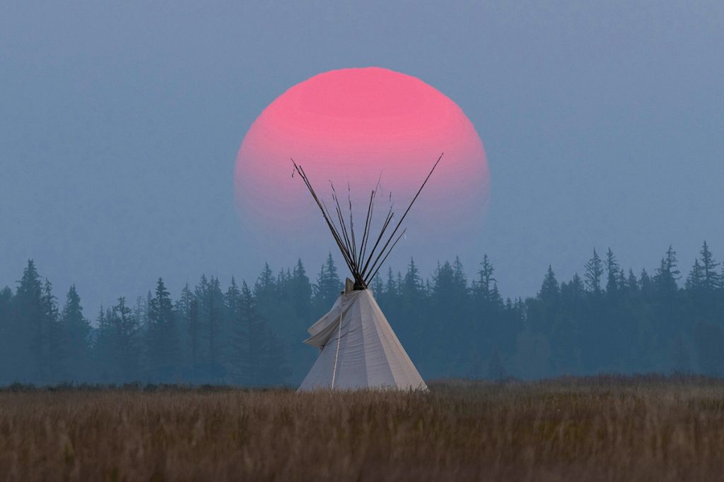A teepee with a large red sun rising behind it.