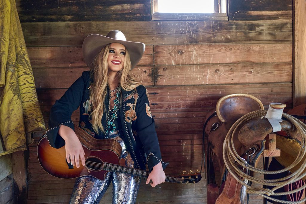 A portrait of Mikayla Lane with her guitar in a tack room.
