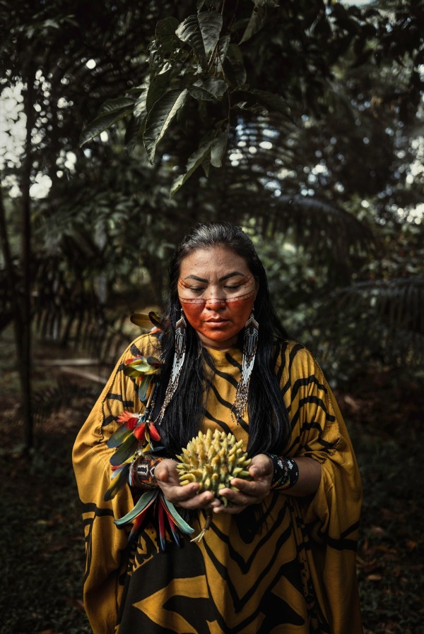 Aldeia Apiwtxa, riserva indigena di Kampa do Rio Amônia: Eliane Yawanawá, moglie del leader Francisco Piyãko, tiene in mano un frutto chiamato biribá. L'abito che indossa si chiama cushma. L'abbigliamento è un elemento centrale nella definizione dell'identità di un popolo. La produzione richiede mesi e puó durare anche un anno. Al giorno d'oggi, gli abiti femminili sono realizzati con tessuti industriali (tocuyo), che vengono peró tinti con colori naturali e poi adornati con dipinti e decorazioni Asháninka dai diversi significati culturali. Eliane appartiene al popolo Yawanawá, è un'artigiana e realizza abiti con motivi della farfalla (yawavana, donna spirituale) e del boa (Runüa), segni di protezione e saggezza ricevuti dal mondo spirituale attraverso la bevanda sacra chiamata kamarãpi. Dalla serie The Forest Knows © Nicoló Lanfranchi
