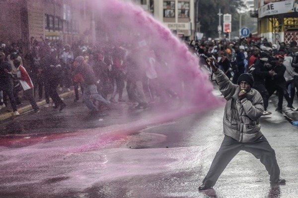 A protestor raises a fist in defiance as Kenyan police officers use a water cannon to disperse the gathering. Pink dye in the water is often used by police at protests to mark individuals for identification. Nairobi, Kenya, 20 June 2024