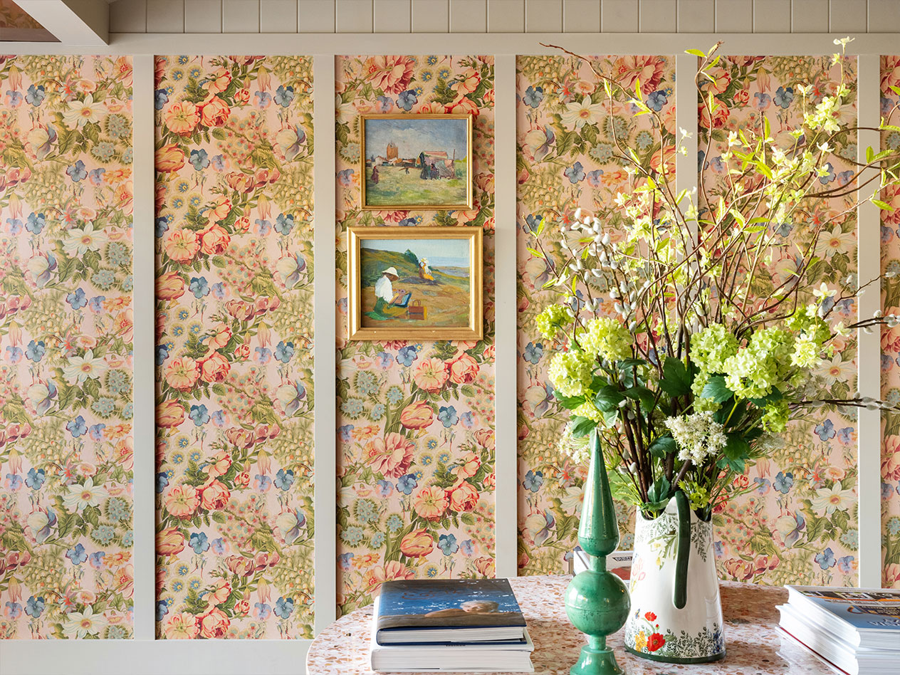 A lobby with floral wallpaper, two framed paintings, a vase with flowers, and books on a marble table.