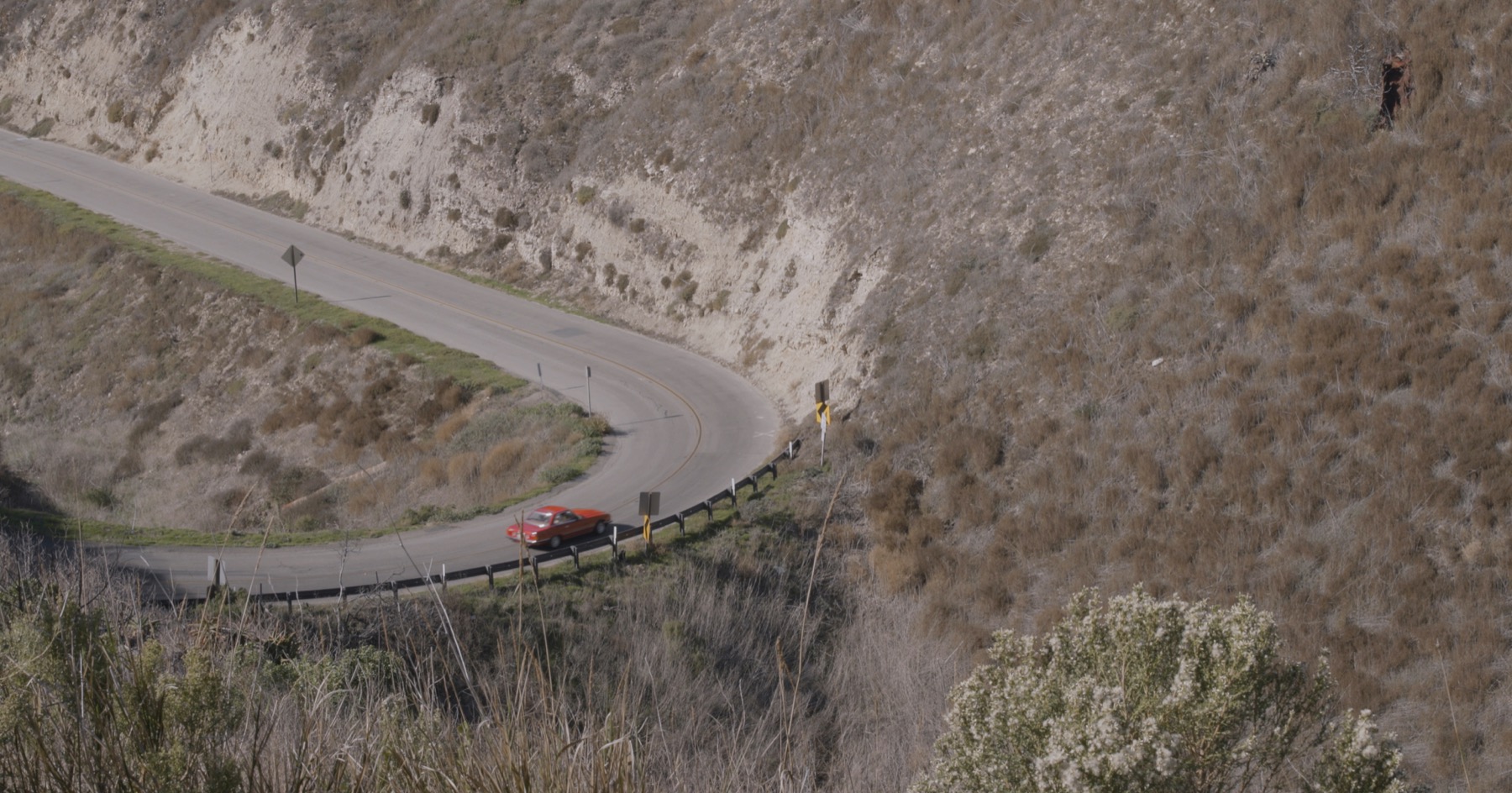 A red car drives on a winding road through a hilly, grassy area.