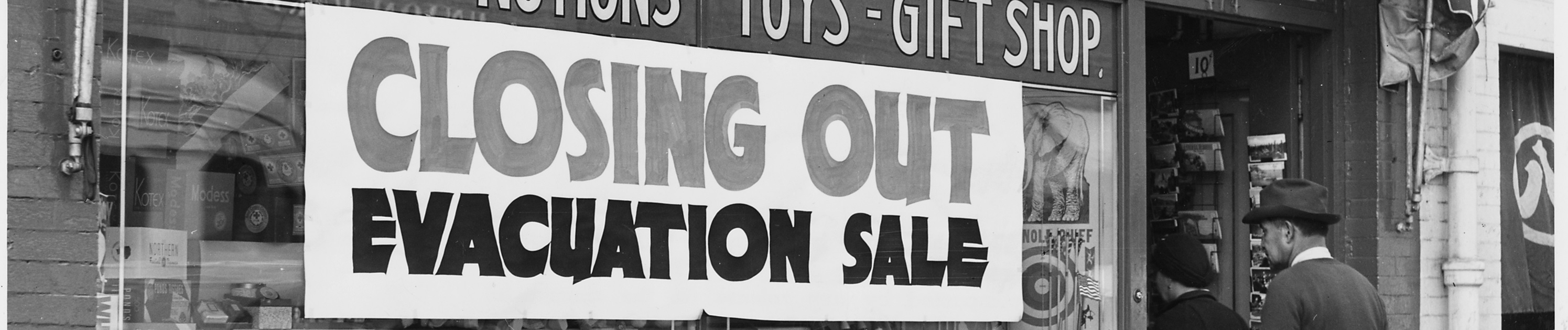 Black and white photograph of a sign in a shop window reading "Closing Out / Evacuation Sale"