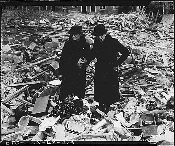 Two ladies in long black coats and black hats standing in the rubble