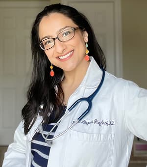 Dr. Krupa Playforth, also known as The Pediatrician Mom, smiling warmly in a white coat with a stethoscope draped over her shoulder, wearing glasses and colorful earrings.