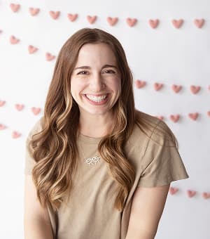 Dr. Taylor Arnold, pediatric dietitian, smiling brightly with long wavy hair, standing in front of a backdrop decorated with small pink heart-shaped garlands.