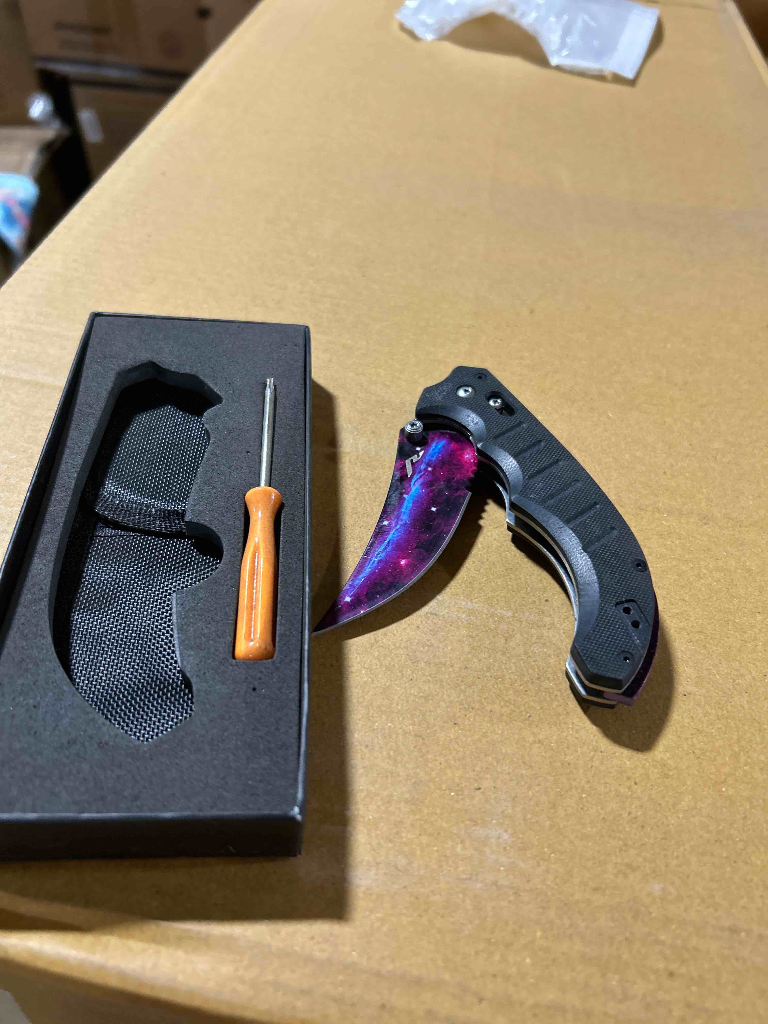 Condition photo showing Brand New for galaxy-finish folding karambit knife set featuring a razor-sharp curved blade, textured ergonomic handle, protective sheath, and adjustment tool, all presented in a premium gift b