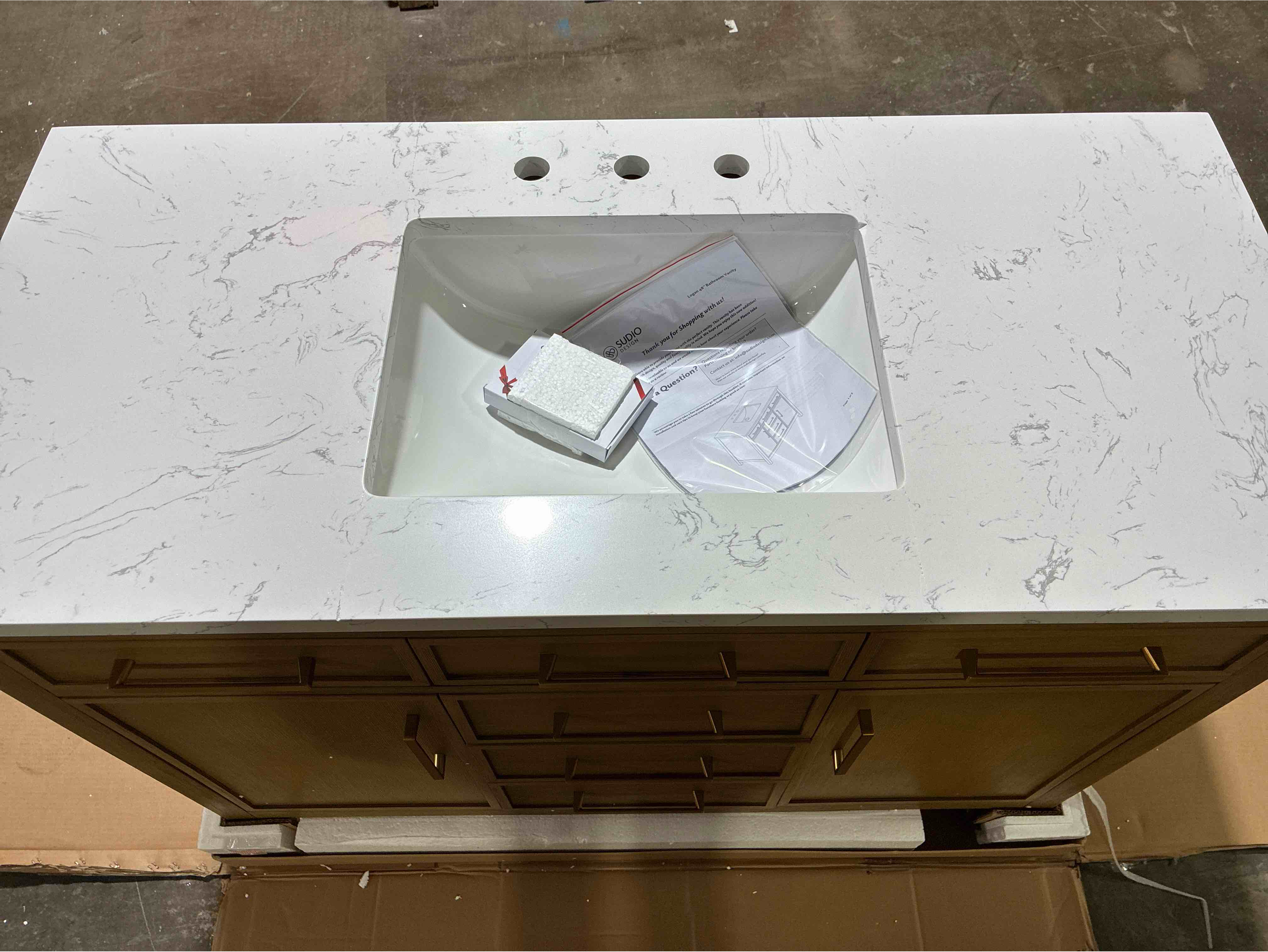 Condition photo showing Good Condition for Reajean 48" Poplar Wood Single Sink Bathroom Vanity with 5 Drawers, 2 Single Shelf Cabinets, Gold Brushed Handles, and Carrara Stone Top. 3 Hairline Cracks Along Sink Edges and Chip along right back rim of sink bowl. Can be coated over sink top remains sturdy. 34'' H X 48'' W X 22'' D