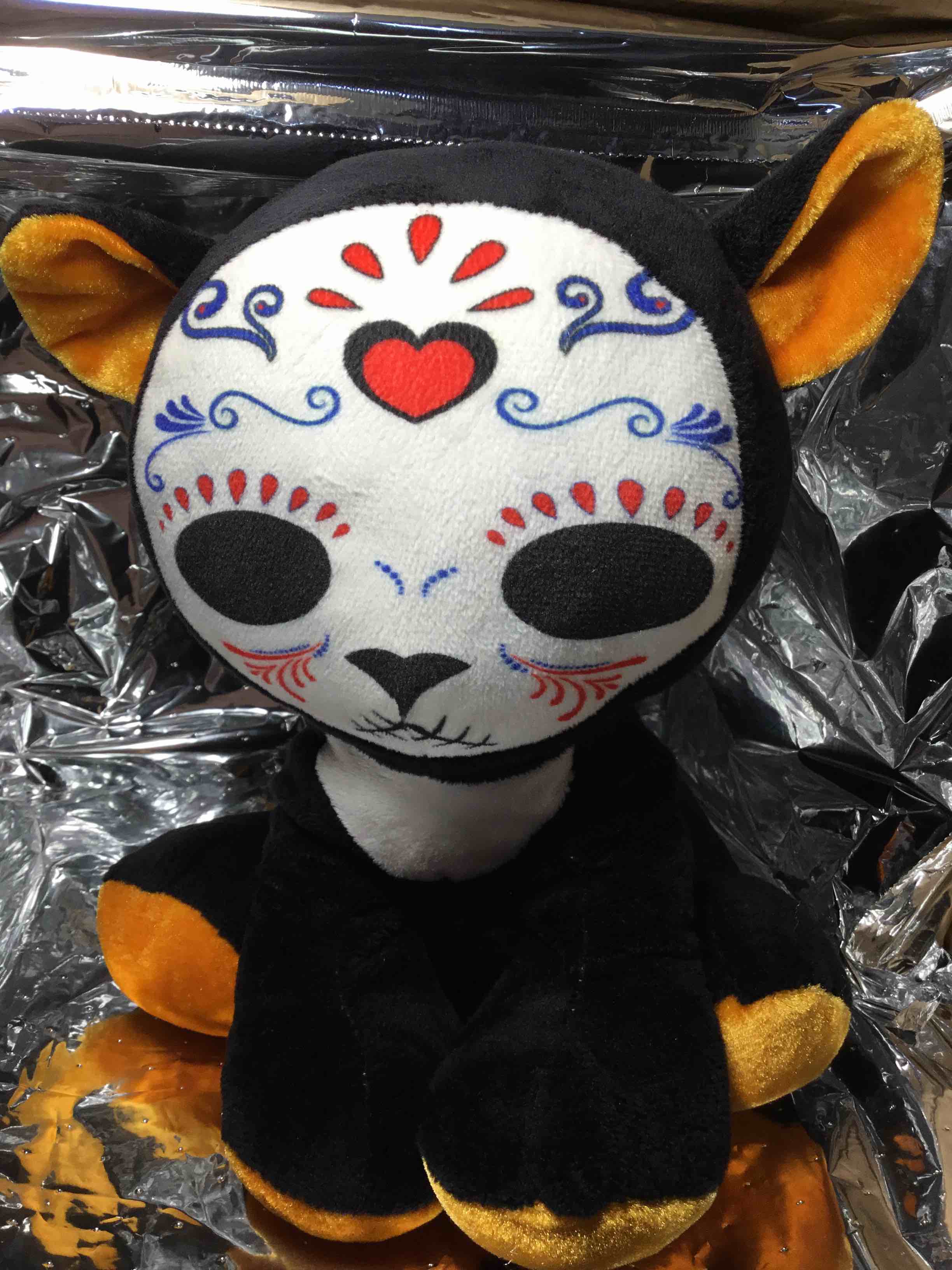 This is a Day of the Dead (Día de los Muertos) themed cat plush. It is a stuffed animal with a sugar skull-inspired design on its face, which is a common symbol of the Mexican holiday
