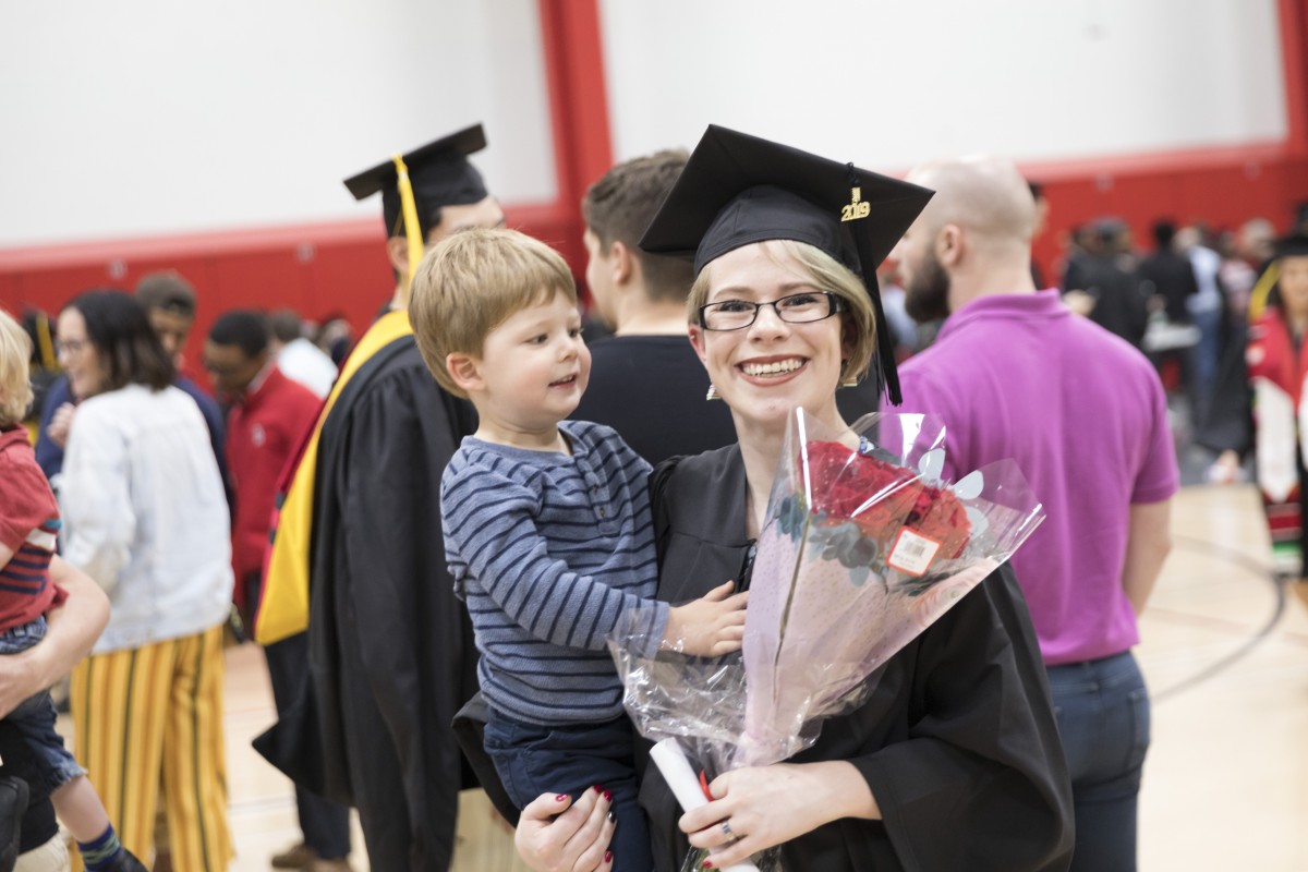 Smiling woman in graduation regalia holding a toddler