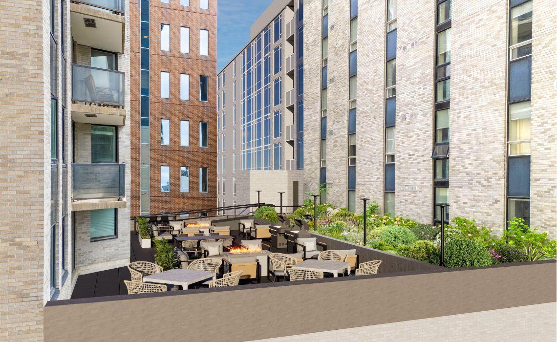 An expansive rooftop patio at The Vantage nestled between modern brick apartment buildings. The space is divided into cozy social nooks with grey outdoor sofas surrounding rectangular fire pits. Lush green privacy plantings, modern black light posts, and m
