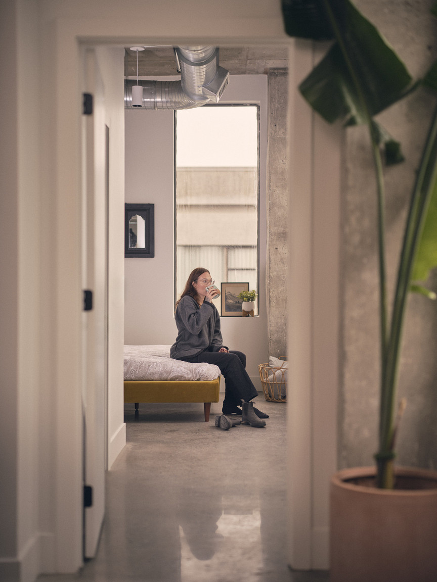 A woman sitting on the edge of a bed drinking a cup of coffee, shot from the hallway looking down at the bedroom.