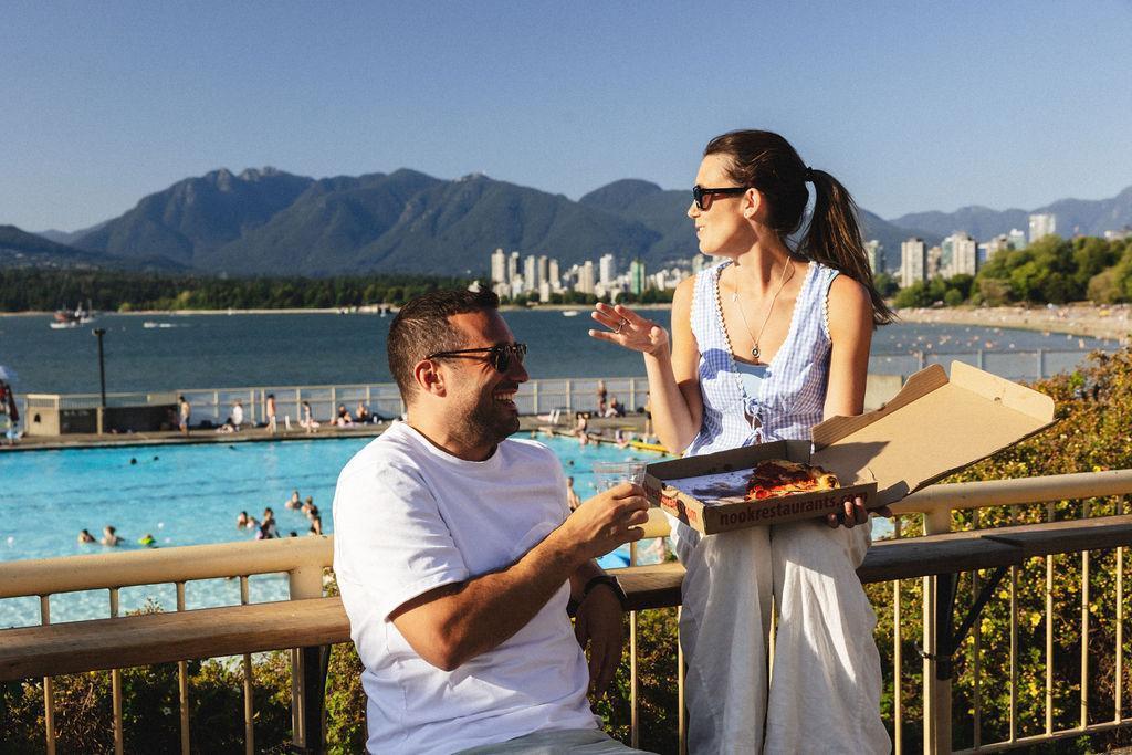 Two people sitting eating pizza, a pool behind them and a mountain range in the background.