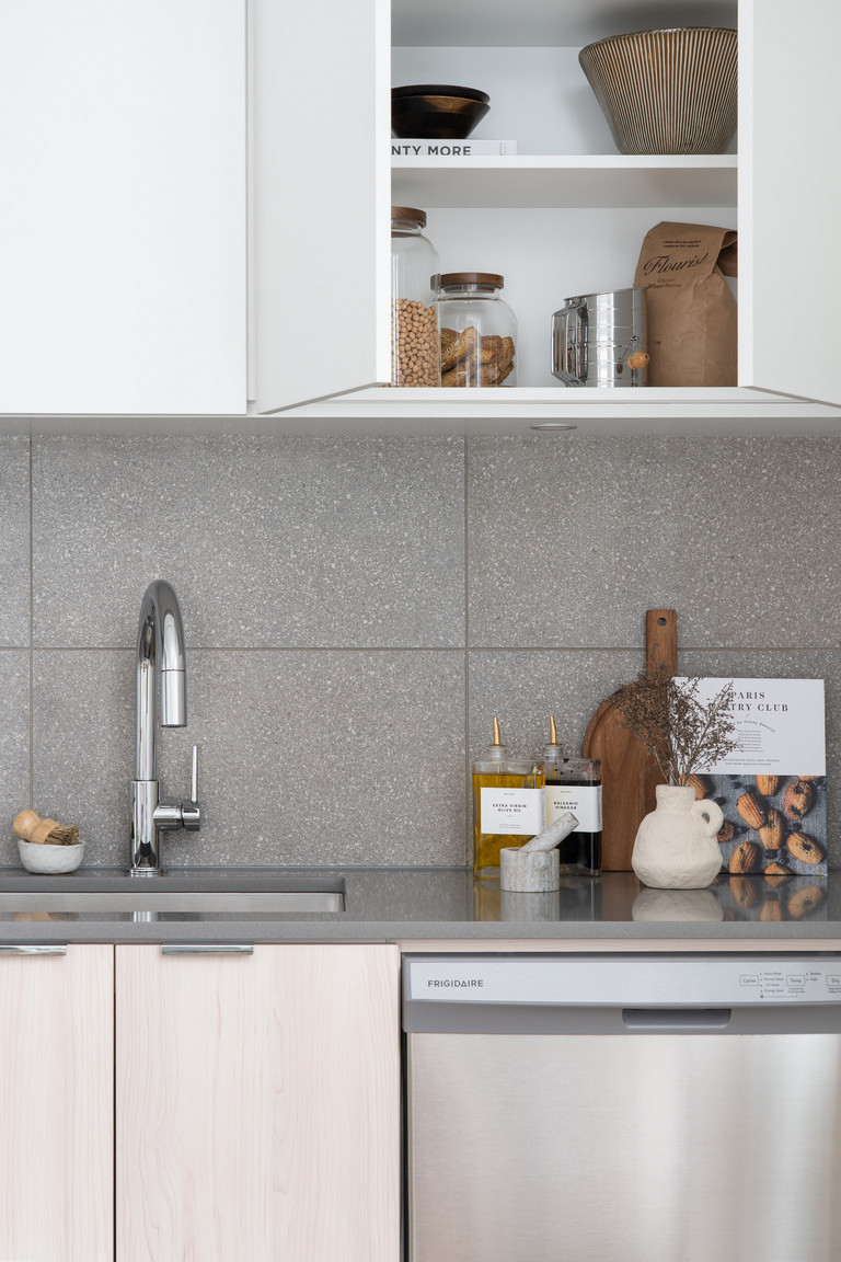 Modern kitchen countertop with stainless steel sink and faucet, open wooden shelves displaying jars, a bowl, and the cookbook 'PLENTY MORE.' Features hand and dish soap bottles, utensil holder, wooden cutting board with dried plants, and an open magazine s