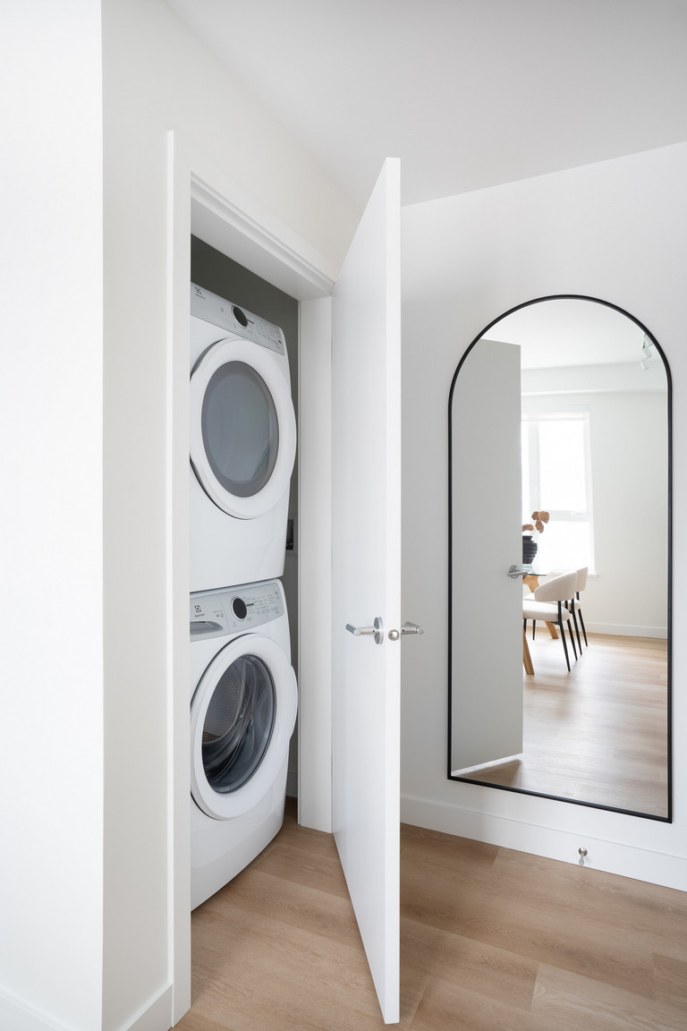 Modern laundry closet with stacked washer and dryer, partially open white door, and adjacent arched wall mirror reflecting a room with wood flooring and contemporary furniture.