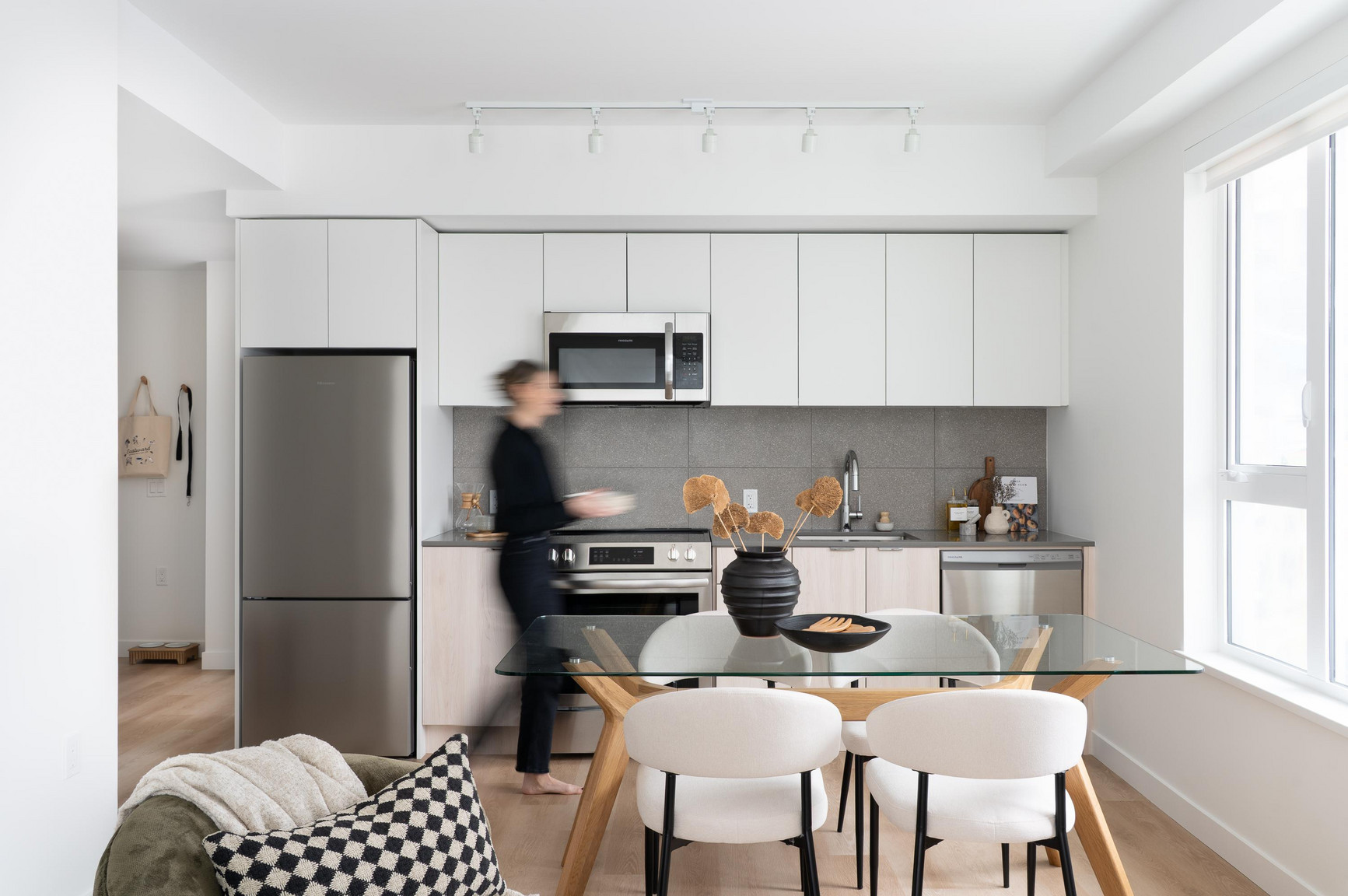 Modern kitchen with white cabinets, stainless steel appliances including refrigerator and built-in microwave, glass dining table with wooden legs and white chairs, black vase with dried flowers, and natural light from large window