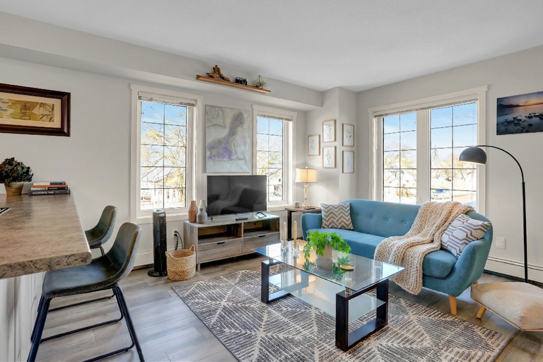 Bright, modern living room featuring a blue mid-century modern sofa, a glass coffee table over a geometric pattern rug, a TV stand, and two bar stools at a kitchen counter next to large windows.