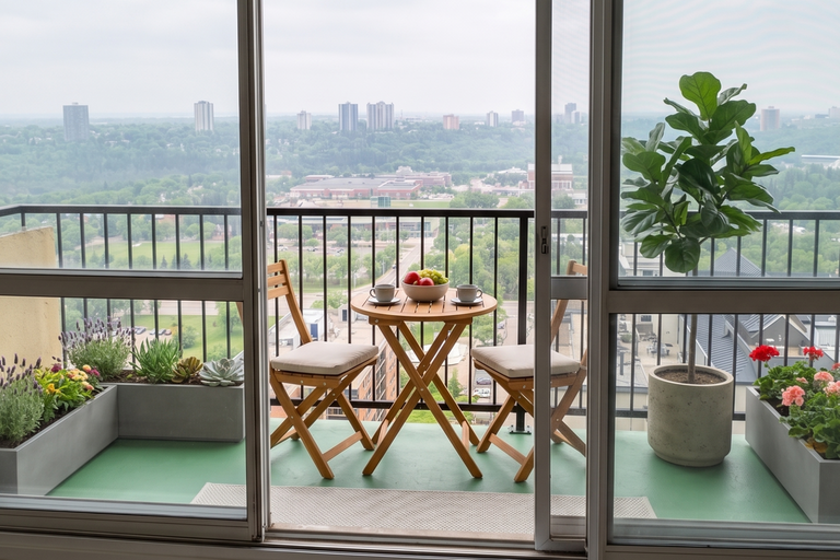 A scenic balcony view from an apartment at Hillside Estate in Edmonton, overlooking a lush green river valley and city skyline. The balcony features a wooden bistro set with fruit and coffee cups, framed by vibrant planter boxes and a fiddle-leaf fig tree.