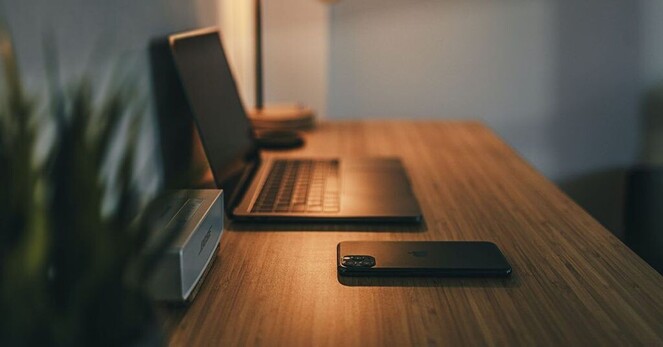 A MacBook sitting on a wooden table with an iPhone face down and dimly lit.
