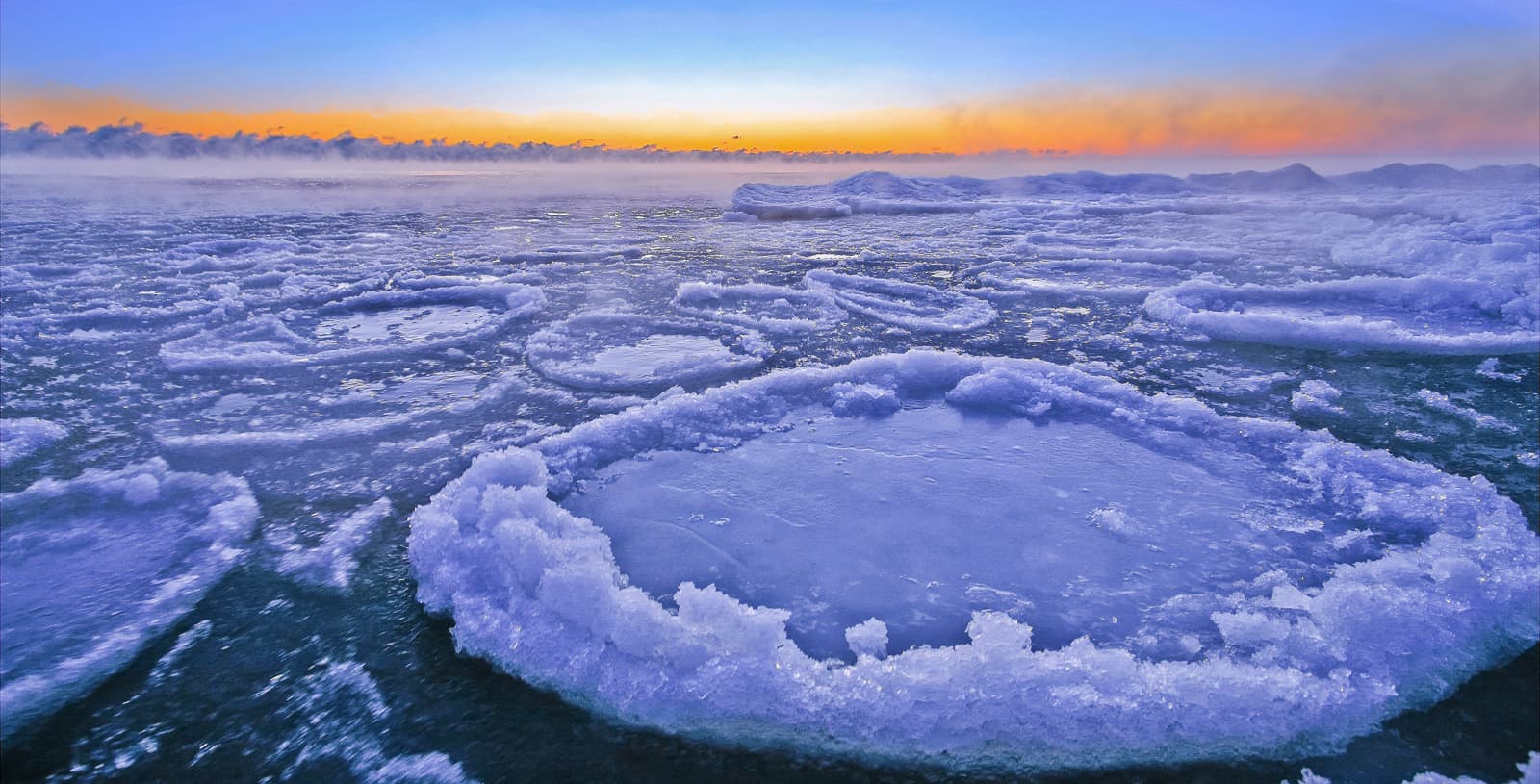 Circular pancake ice with jagged edges and malformed chunks floating in mist on a lake at sunset.