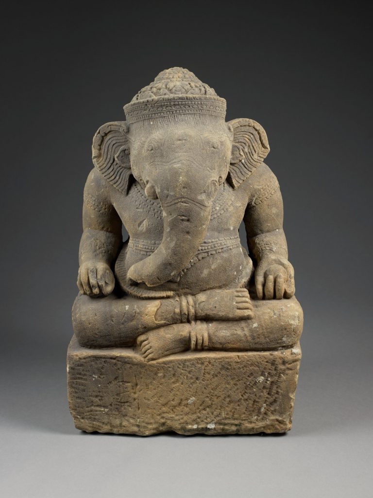 Photograph of a sculpture depicting Ganesha, the Hindu deity with an elephant head. They are sitting crossed-legged on a mall platform.