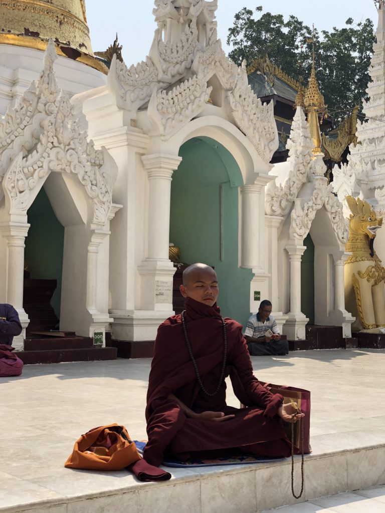 Photograph of a monk meditating in front of a white and gold temple. He is wearing a dark garment covering most of his body and holding prayer beads; his eyes are closed.