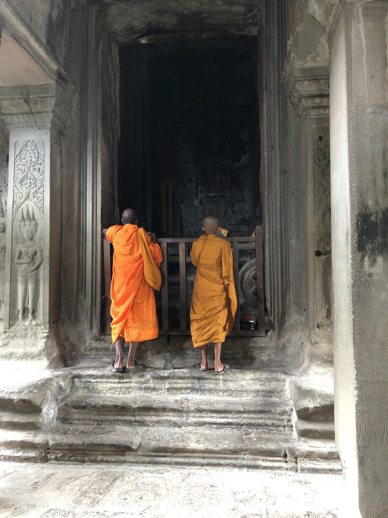 Photograph of two monks dressed in orange garments. They are standing with their backs to the camera, looking at details of a shrine