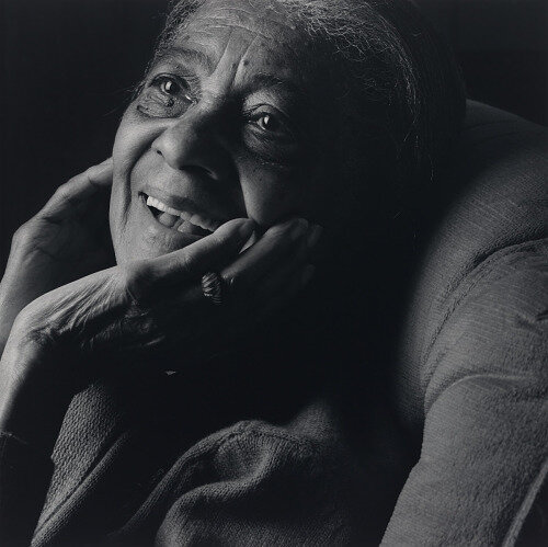 The photo accompanying the title slide for Path 2 is a black-and-white portrait of Elizabeth Cotten on the right side of the slide. Depicting her in her more advanced years, her head rests against a cushion, her chin rests in her hand with a happy smile on her face. She looks off to the upper left corner of the frame.