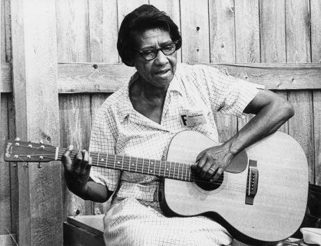 This black and white front-view photo of Elizabeth Cotten, on the right side of the slide, again shows her unusual style of holding the guitar, strumming the strings with her left hand and fingering the notes with her right. She sits outside in front of a wooden fence, wearing glasses.