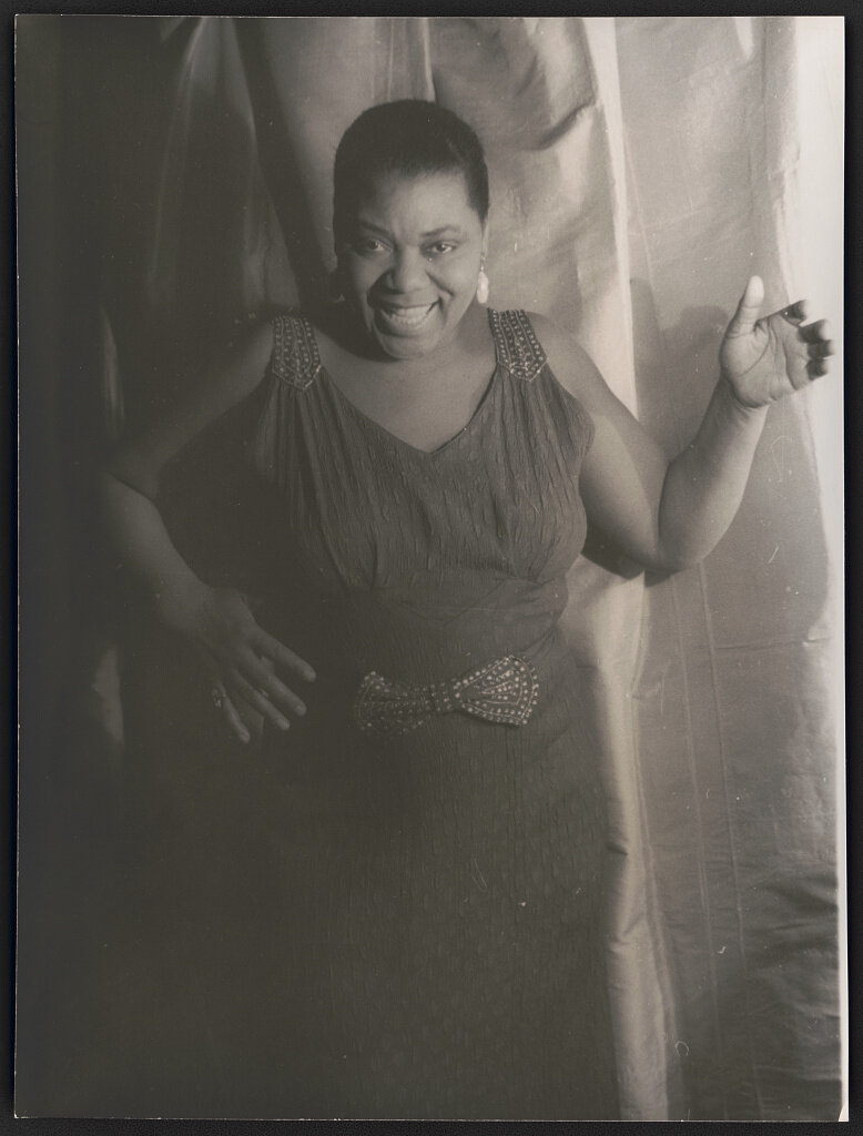 This ¾ length photograph of Bessie Smith shows her at a later stage in her career. She stands smiling in front of a light drapery, with her left hand raised and her right hand resting on her waist. She wears a sleeveless dress that has a cloth bow at the waist and matching shoulder straps. Her hair is very short.