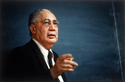This color photograph of Sterling Brown shows him formally dressed in the typical style of a university professor delivering a lecture. He wears a dark suit, white shirt, red tie. He holds up his right hand with his finger outstretched as if to make a point.