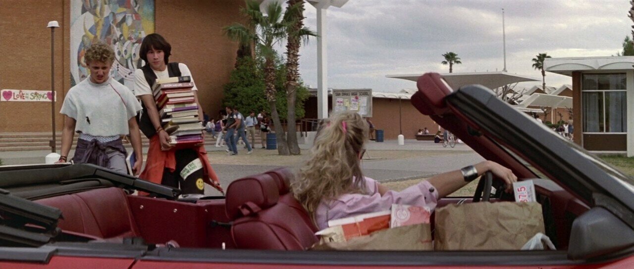 Bill and Ted walk towards a convertible car with Bill's stepmom in the drivers seat. Ted is holding an enormous stack of books