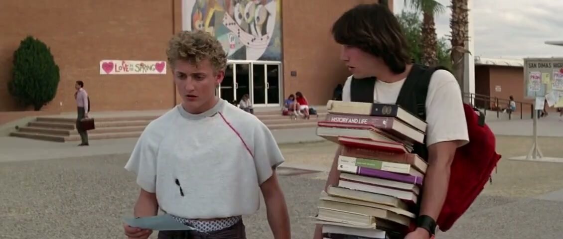 Bill and Ted look look serious, standing outside their school. Ted has a huge stack of history books in his arms