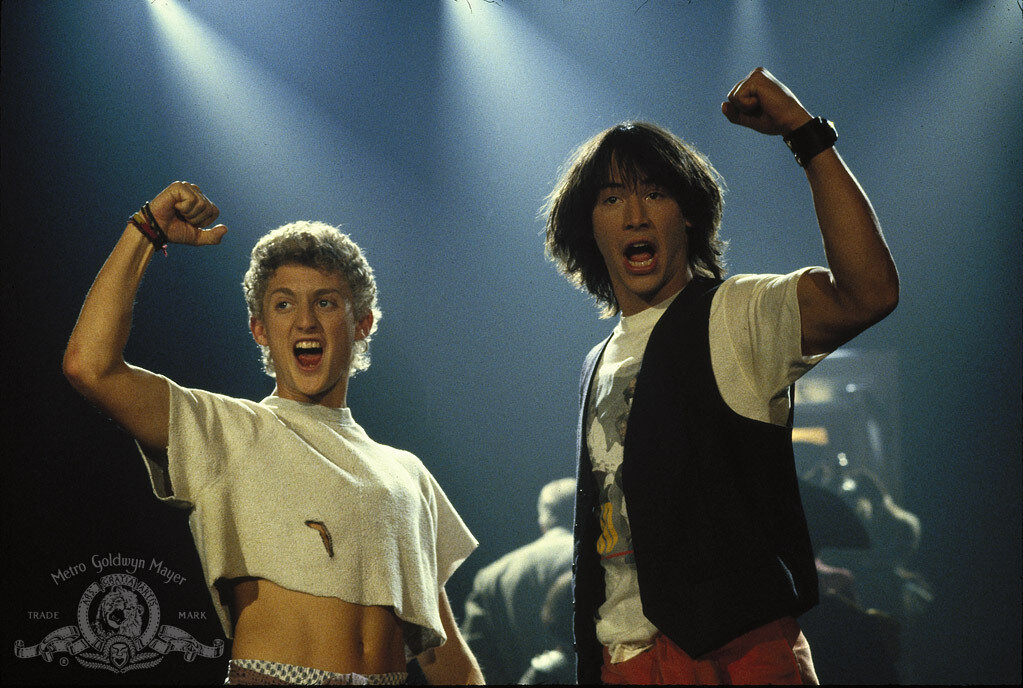 Bill and Ted look triumphant, fist raised in the air with spotlights from above. Most triumphant! Bill is wearing a crop top exposing some belly, and Ted is in his standard waistcoat