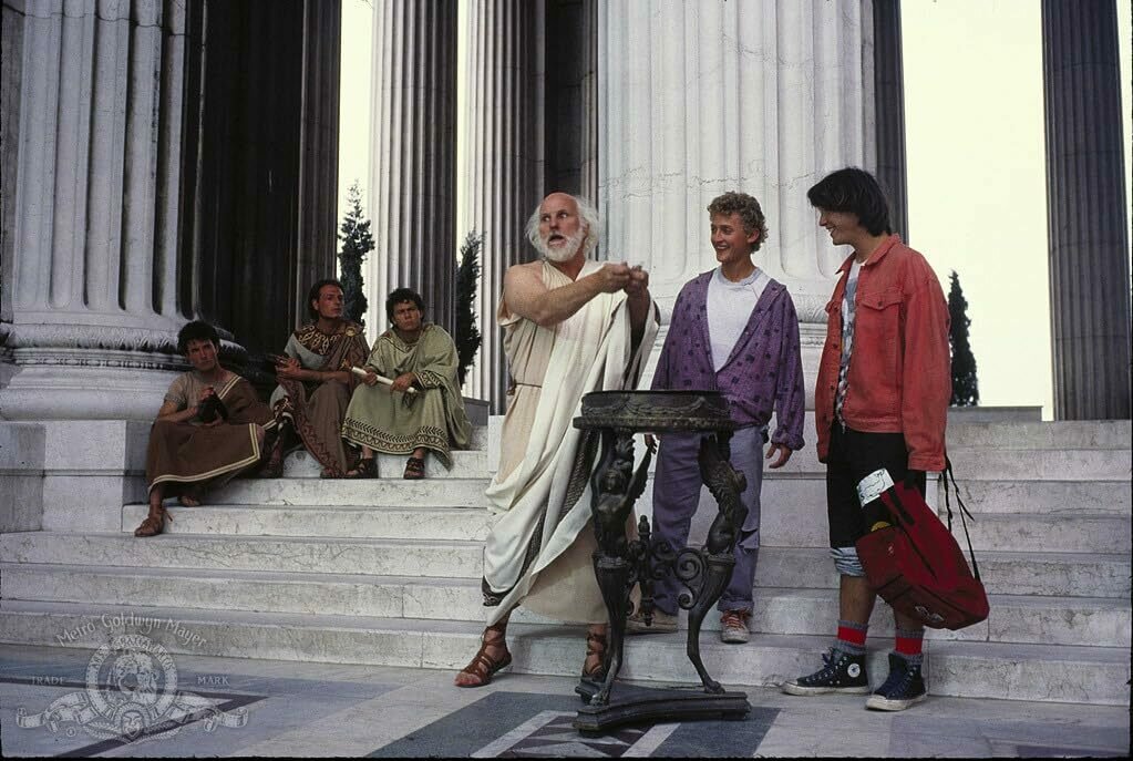 Bill and Ted philosophise wiht Socrates in front of ancient greek columns. They look amused, Socrates serious