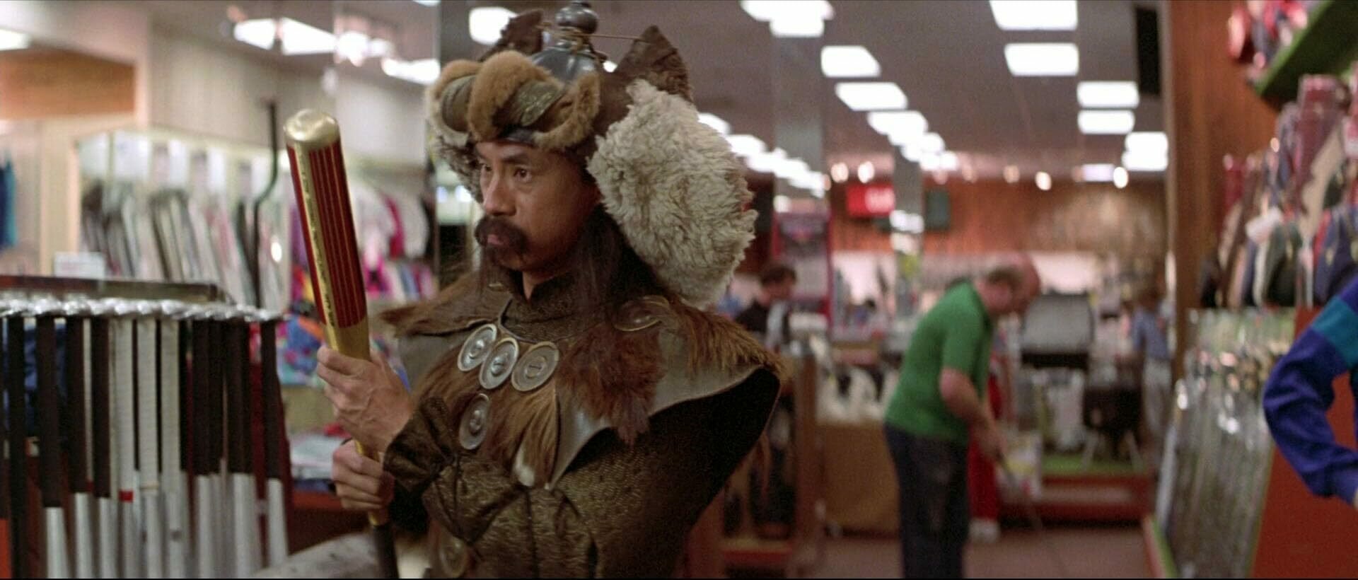 Genghis Khan looks at a baseball bat in a sport shop. He's wearing lots of furs and is looking at the bat with great interest