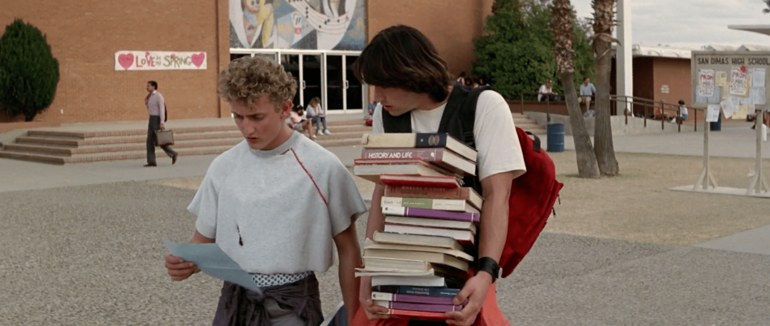 Bill and Ted walk towards a convertible car with Bill's stepmom in the drivers seat. Ted is holding an enormous stack of books