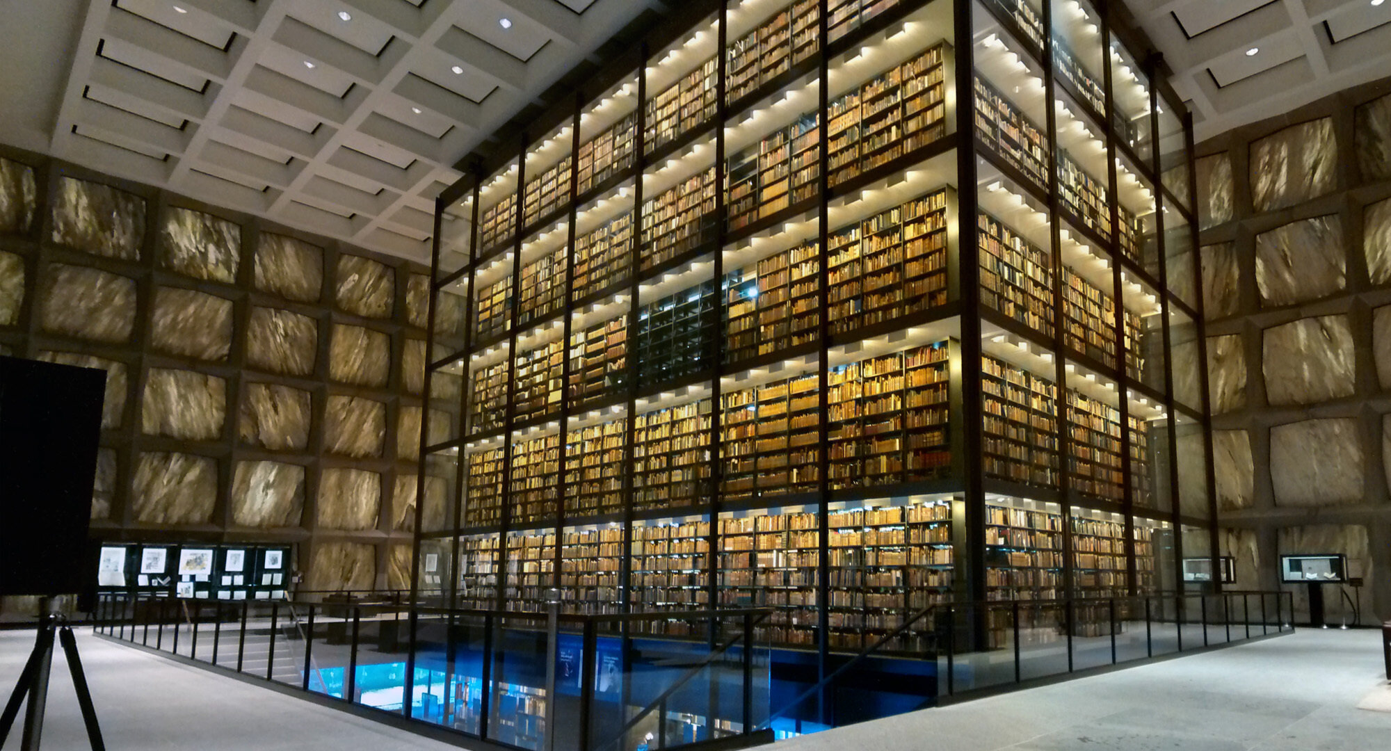 Interior de la Beinecke Rare Book &amp; Manuscript Library en la Universidad de Yale, en New Haven, Connecticut. Se aprecia una extensa colección de libros raros y manuscritos almacenados en estanterías de vidrio que alcanzan desde el suelo hasta el techo. La iluminación cálida destaca los lomos de los libros y crea un ambiente sereno. La estructura arquitectónica de la biblioteca, con paredes de mármol translúcido, permite que la luz natural se filtre suavemente en el espacio, protegiendo los valiosos materiales de la exposición directa a la luz. La biblioteca, conocida por su diseño único y sus impresionantes colecciones, es un punto de interés tanto para investigadores como para visitantes interesados en la historia del libro y la erudición.