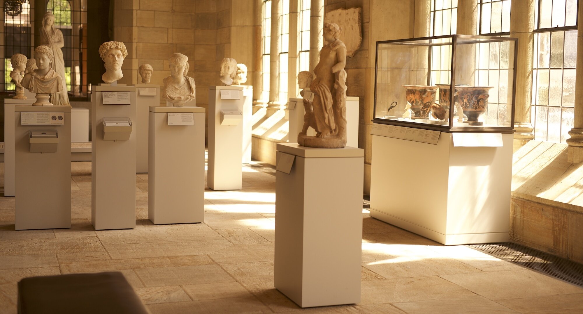 Sala de la Yale University Art Gallery, bañada por la luz natural que se filtra a través de las ventanas altas. Se observan varias estatuas clásicas y bustos en pedestales, creando un ambiente tranquilo y contemplativo. Cada obra está acompañada por su propia etiqueta descriptiva, permitiendo a los visitantes aprender sobre su historia y significado. A la derecha, se ve una vitrina que alberga vasijas antiguas, destacadas por la luz que entra por la ventana y refleja sus detalles intrincados. La exposición combina elementos de la arquitectura clásica con una disposición moderna y accesible, invitando a la reflexión y al estudio de las piezas.