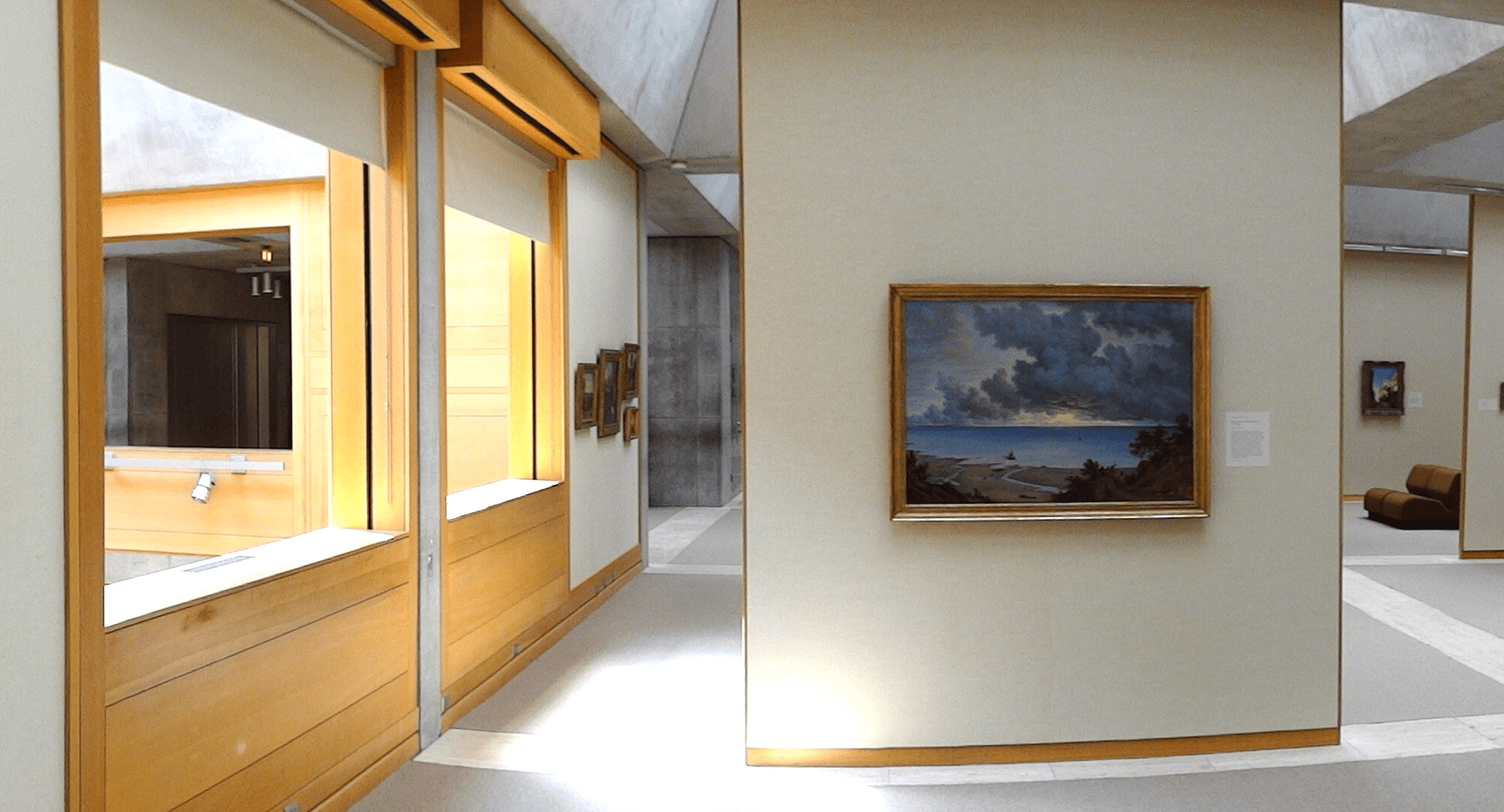 Interior del Yale Center for British Art, con una decoración minimalista y moderna. A la izquierda, se observan ventanas que dejan entrar luz natural y exhiben algunas obras de arte enmarcadas. En el centro y a la derecha, se destaca una pintura de un paisaje marino colgada en la pared, con colores oscuros que resaltan la dramática escena de nubes y cielo en una playa. El espacio es amplio y acogedor, con sofás marrones que invitan a los visitantes a detenerse y contemplar las obras. La arquitectura combina elementos de madera clara y detalles en concreto, creando una atmósfera de tranquilidad y reflexión.
