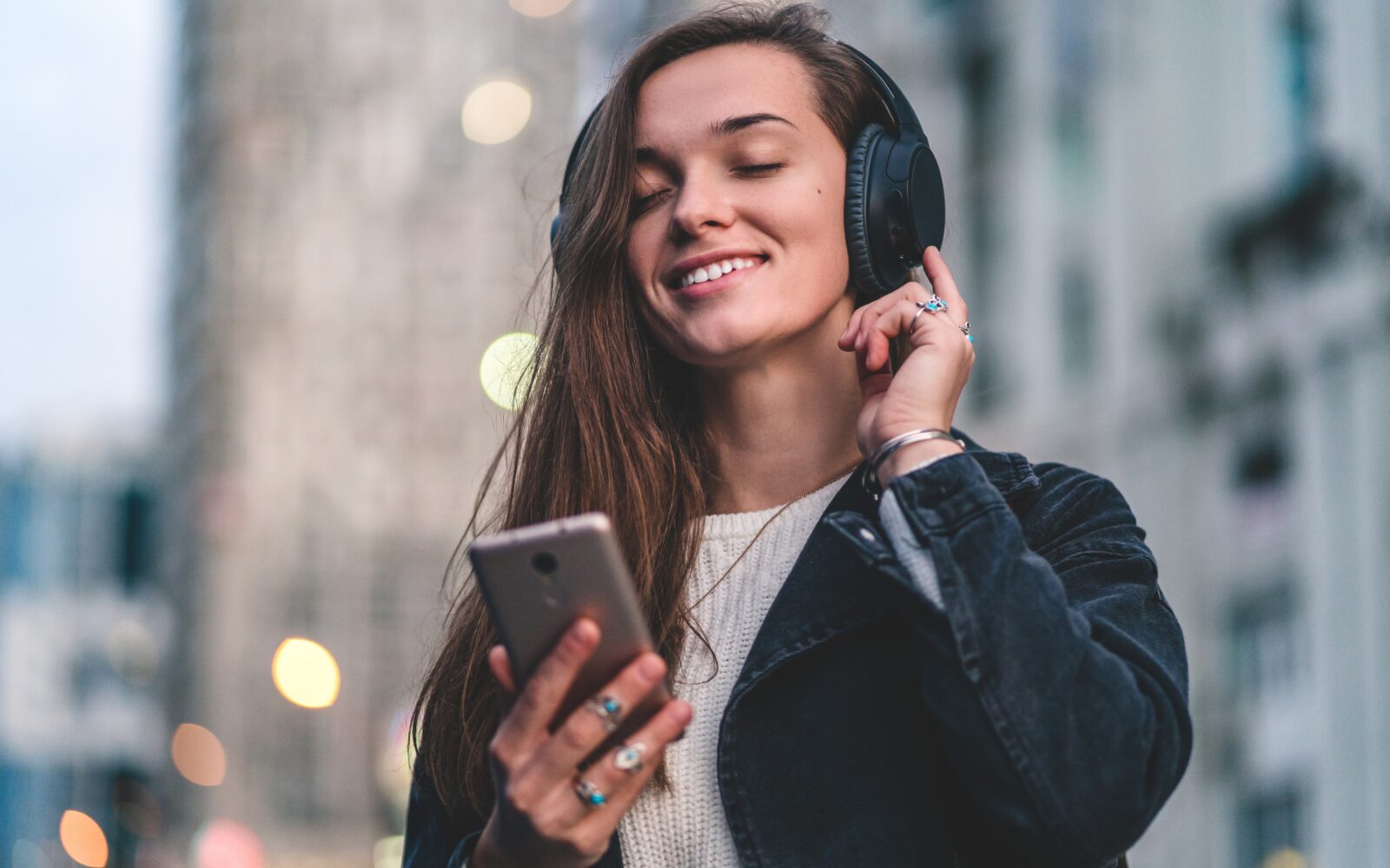 Young happy stylish trendy casual hipster woman changes songs and tracks on smartphone during listening to music on a wireless headphone while walking around the city Music lover enjoying music