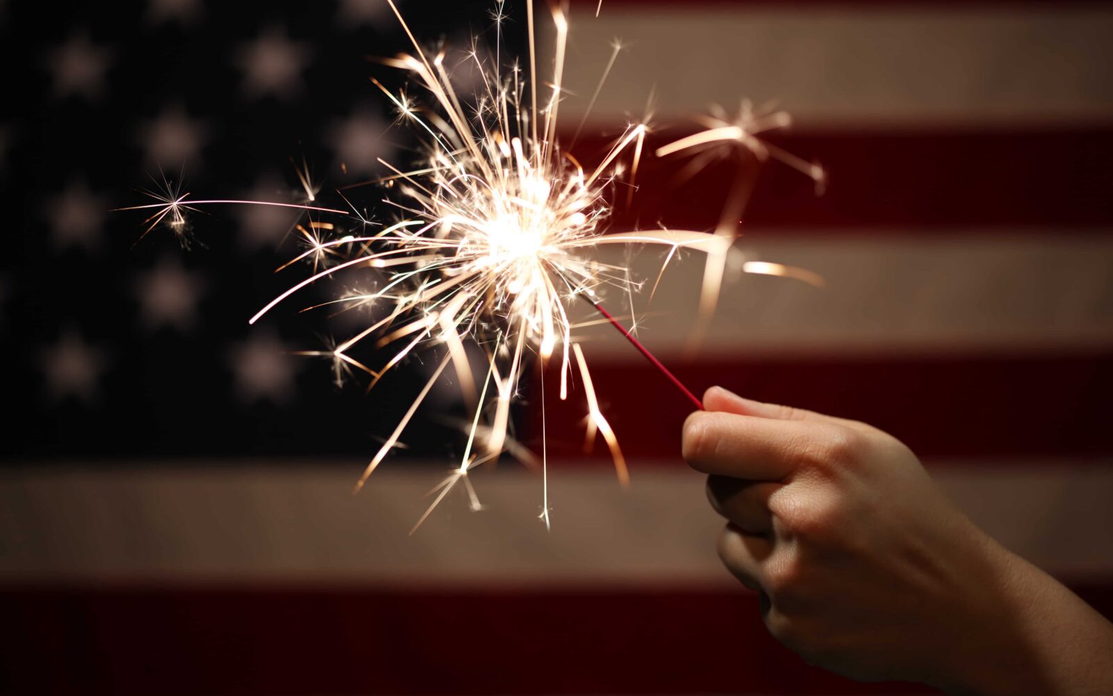 Hand holding lit sparkler in front of the American Flag for 4th of July celebration