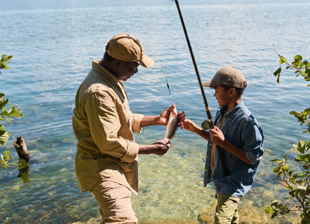 happy mature black man in activewear showing fish to his grandson with fishingrod