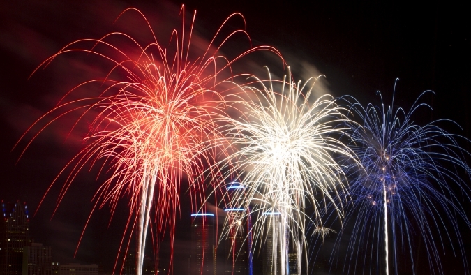 Red, white, and blue fireworks bursting over a city skyline at night during a July celebration.