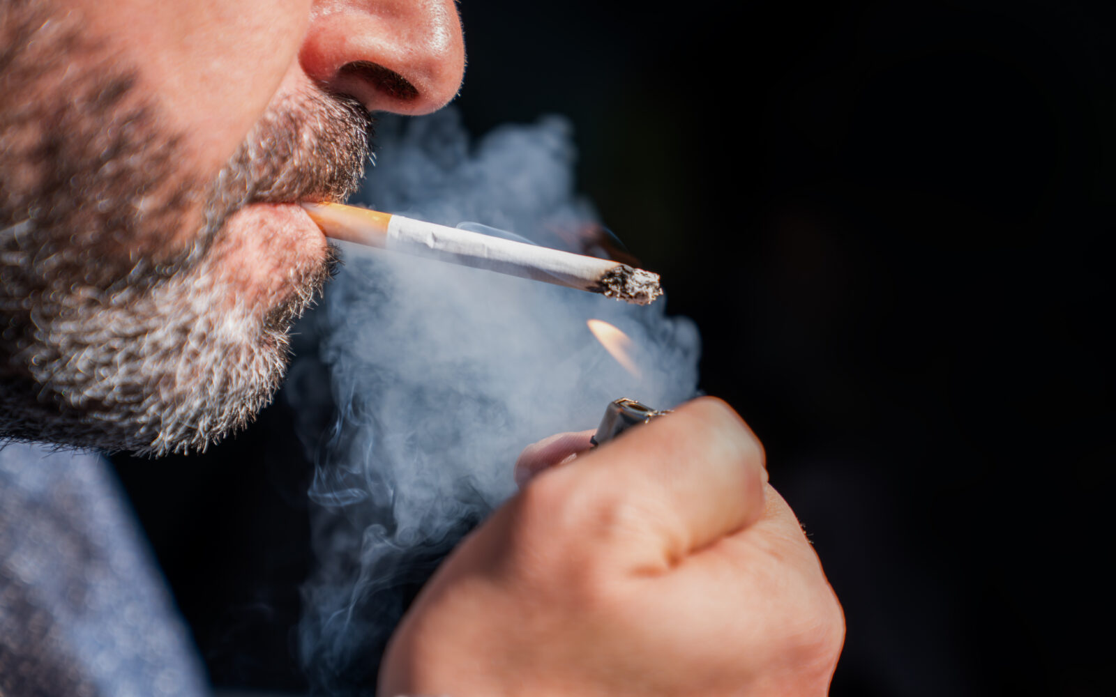 Close up of bearded man holding a lighter and smoking a cigarette on black background