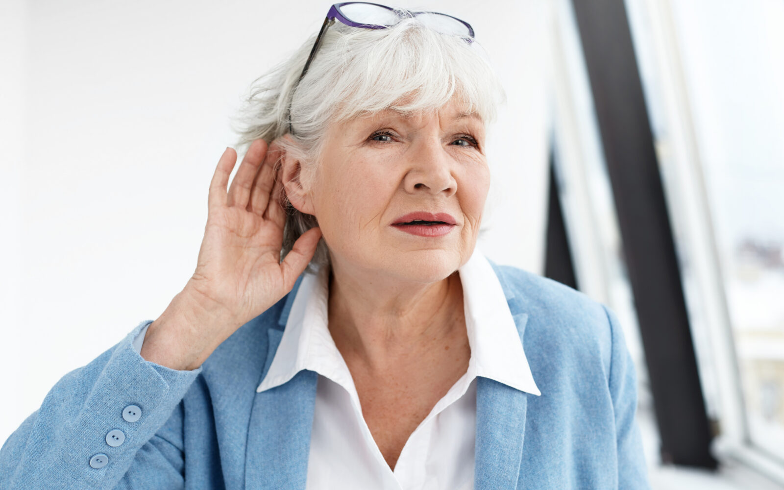 Come again please? Attractive mature woman wearing blue jacket and spectacles on her head eavesdropping Stylish retired female in elegant clothes holding hand at her ear, suffering from hearing loss
