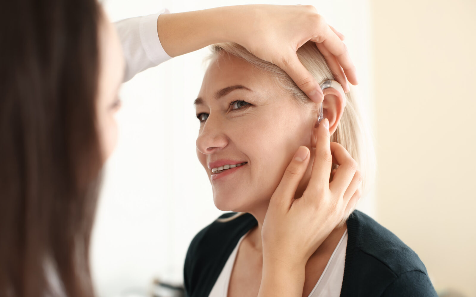 Otolaryngologist putting hearing aid in woman's ear on light bac