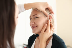 Otolaryngologist putting hearing aid in woman's ear on light bac
