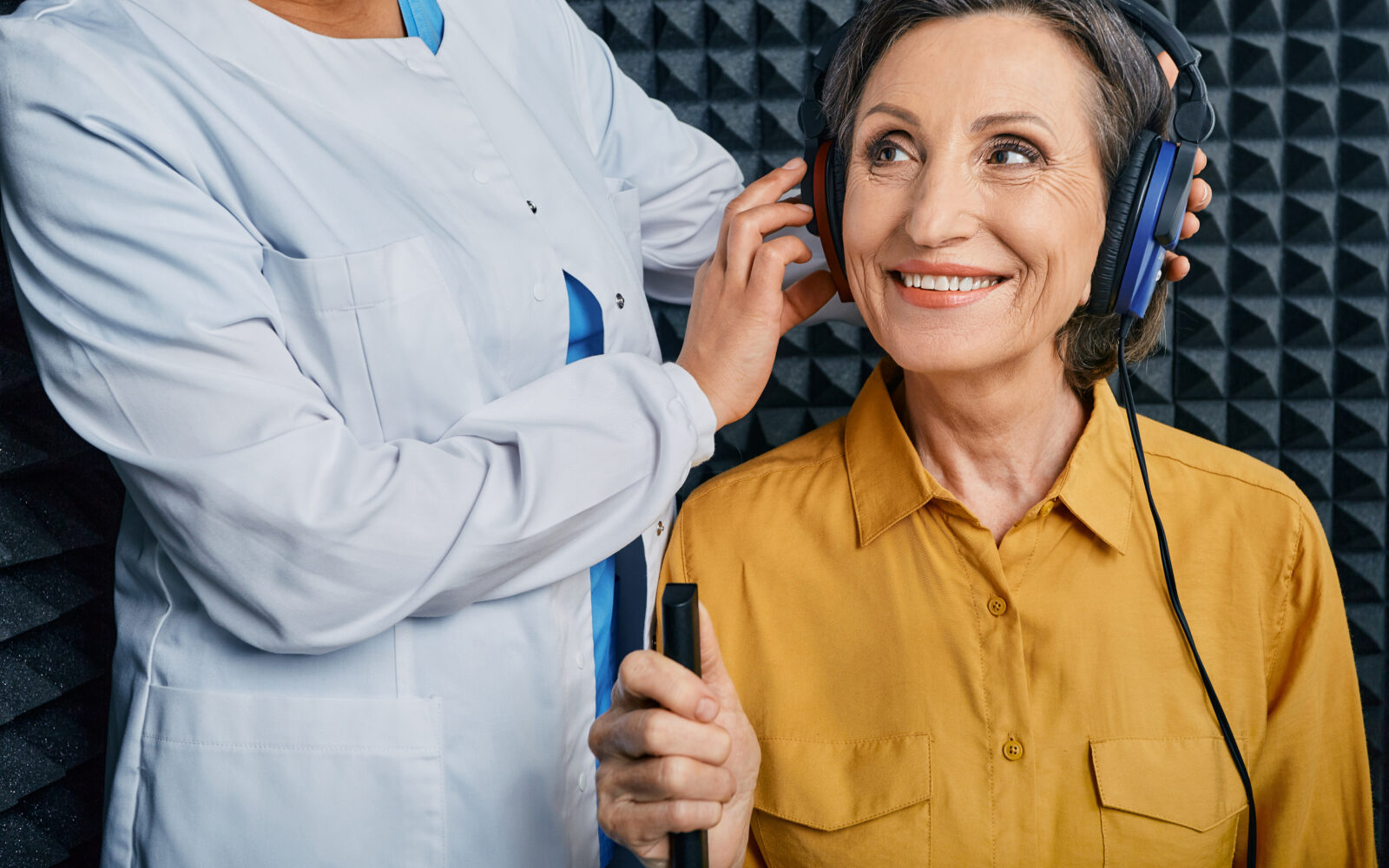 Portrait senior woman with white toothy smile while hearing check up with ENT doctor at soundproof audiometric booth using audiometry headphones and audiometer