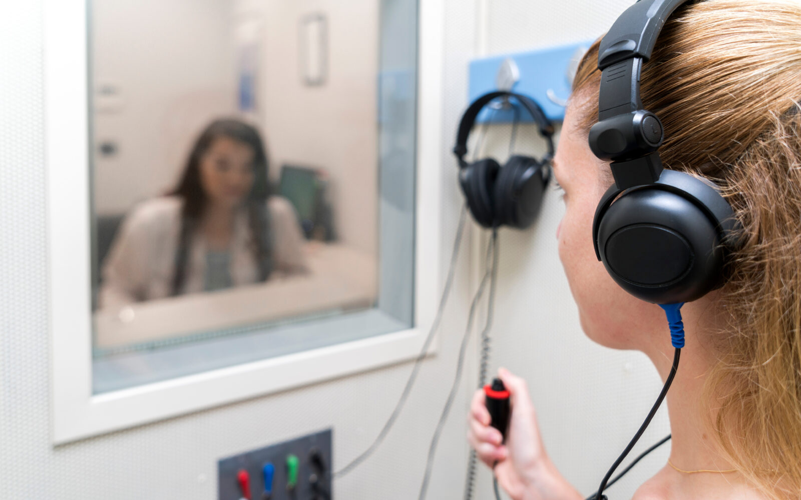 A woman wearing headphones holds a response button during a hearing test in a soundproof booth, with an audiologist visible through the window.